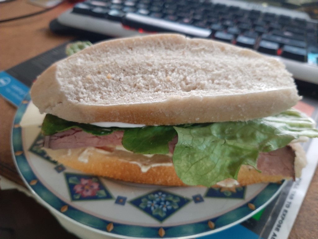 A roast beef sandwich with lettuce and tomato and mayo on a plate. In the background is a keyboard.