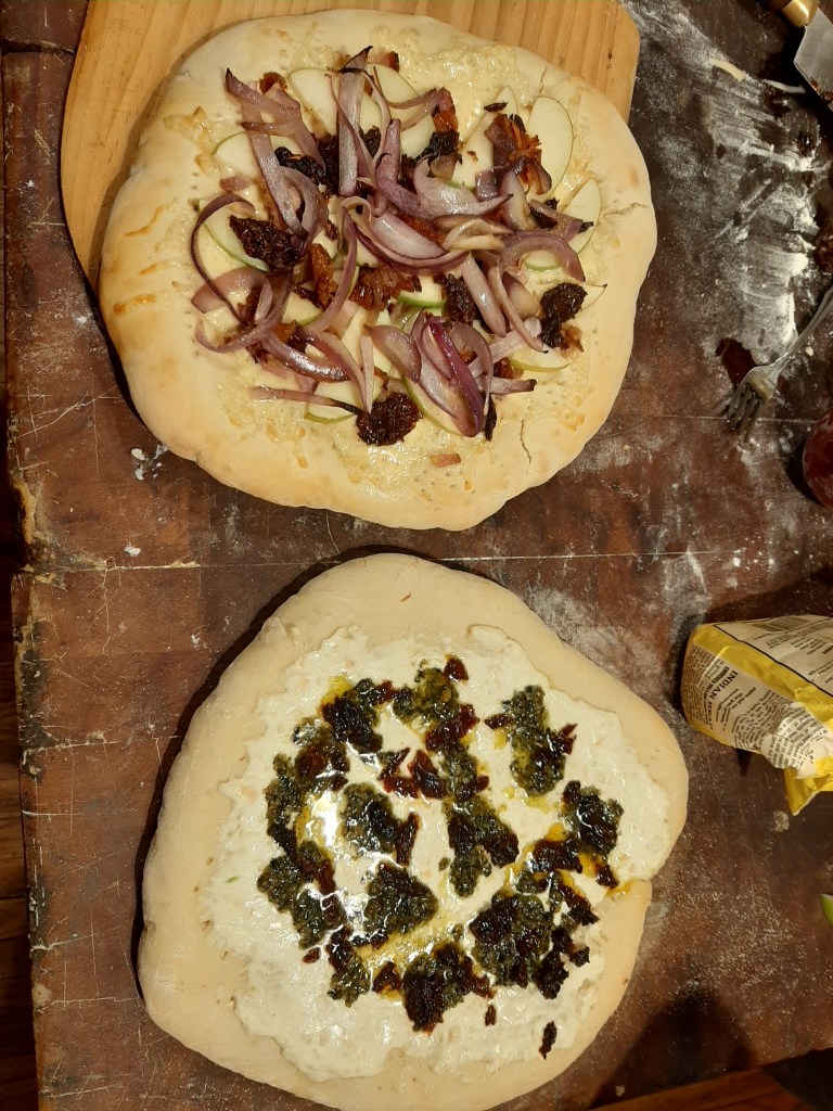 Image of two pizzas looking from above. On top of a butcher block.
Top one has fried onions, Granny Smith apple, bacon, sundried tomatoes.

Bottom one is white pizza with home made pesto.
