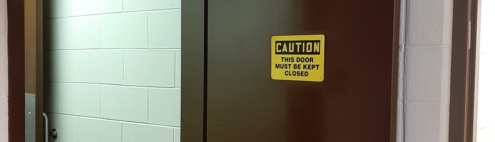 Yellow sign with black lettering on propped open door. Sign says "Caution This door must be kept closed"